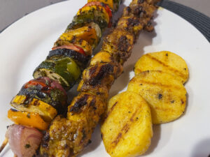Grilled kebab skewers with charred chicken and colorful peppers beside golden, spice-dusted polenta slices on a white plate; a hearty, appetizing meal.