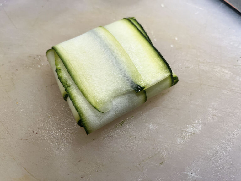 A neat square parcel of overlapping pale-green zucchini ribbons, edged with dark green peel, rests on a cutting board, suggesting fresh, careful preparation.