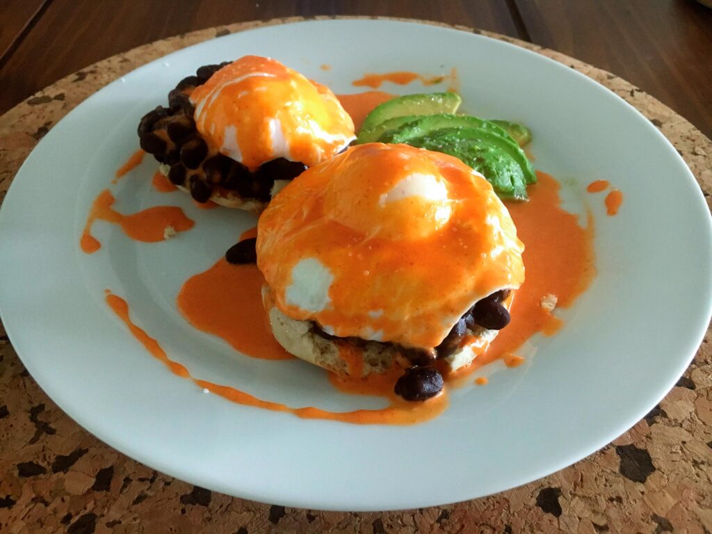 Two English muffin halves topped with black beans, poached eggs, and orange sauce on a white plate, with sliced avocado; a hearty, casual meal.
