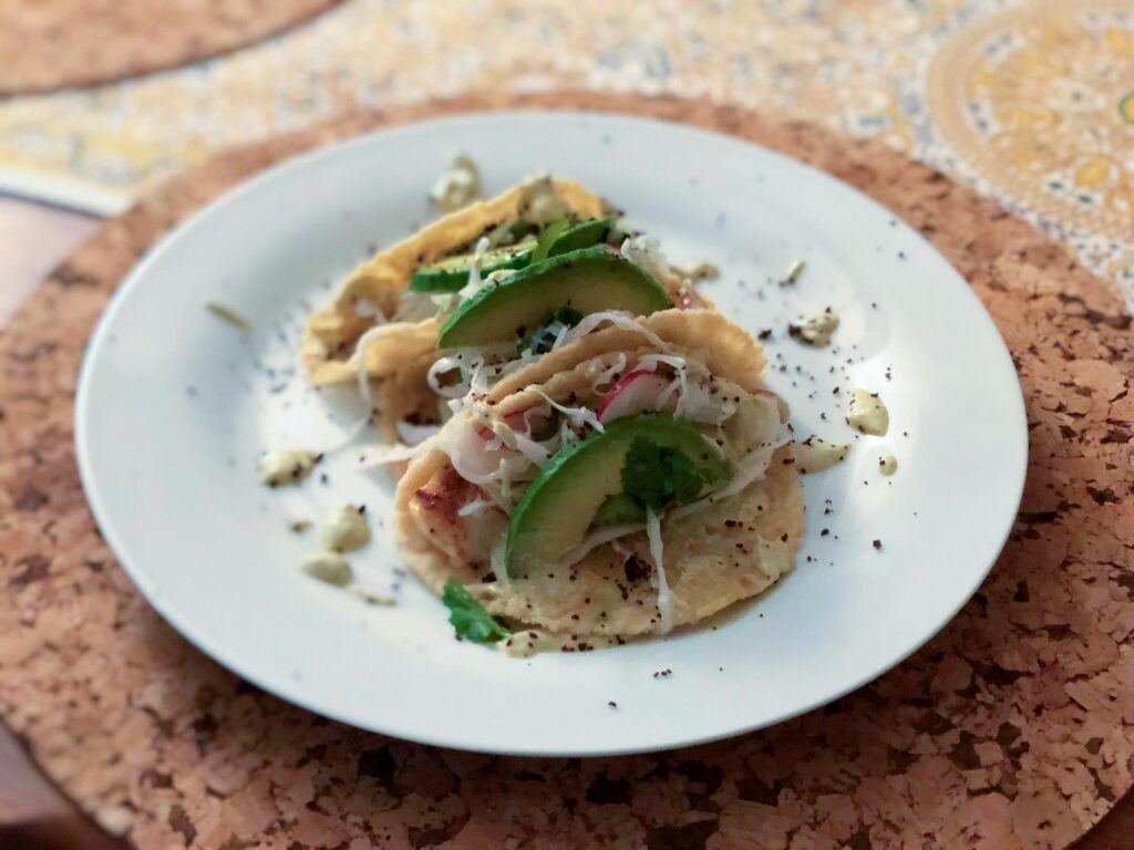Two soft tacos filled with fish and vegetables, topped with avocado slices and herbs, sit on a white plate; a warm, rustic meal.