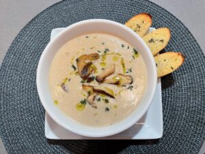 A white bowl of creamy mushroom soup, drizzled with olive oil and herbs, sits on a square plate with three toasted bread slices; warm, inviting presentation.