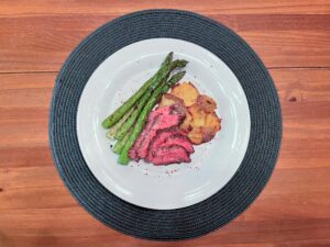 A white plate on a dark woven placemat holds sliced pink-centered roast beef, grilled asparagus, and golden roasted potatoes on a wooden table, presenting a warm meal.