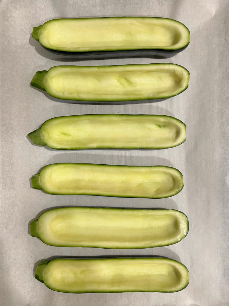 Six lengthwise-cut zucchini halves arranged in a vertical column on a pale surface, showing pale yellow-green flesh and dark green skin; a simple, orderly food-preparation scene.