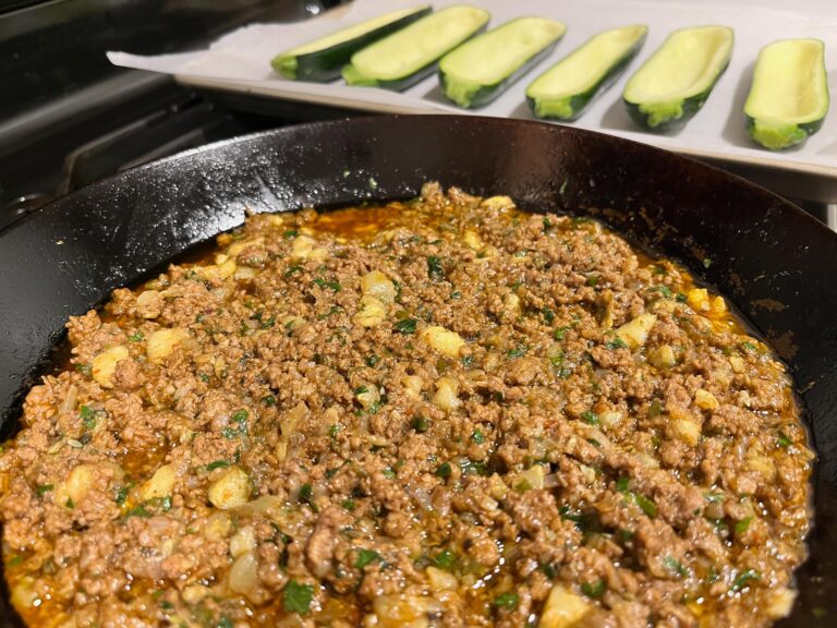 A black skillet holds a browned ground-meat and potato mixture flecked with herbs, while sliced zucchini rests on a tray behind it, ready to cook.