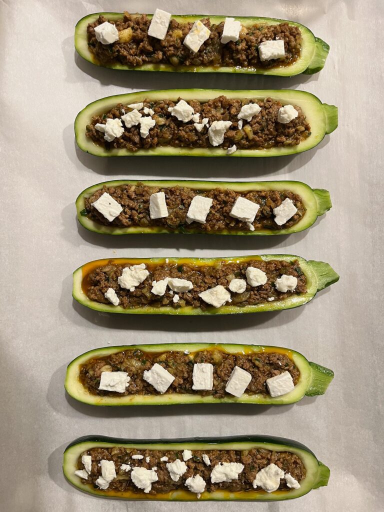Six halved zucchini boats are lined up on a white surface, filled with browned minced meat and topped with chunks of white cheese; appetizing, homemade food.