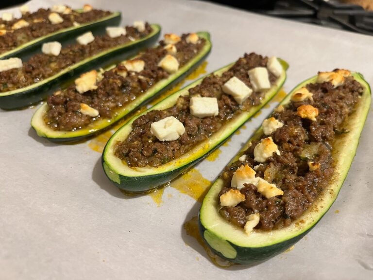 Several zucchini boats are filled with browned minced meat and cheese, arranged on a baking tray; the golden, hearty dish looks warm and appetizing.